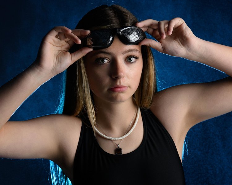 Studio portrait of a young swimmer adjusting her goggles against a blue backdrop